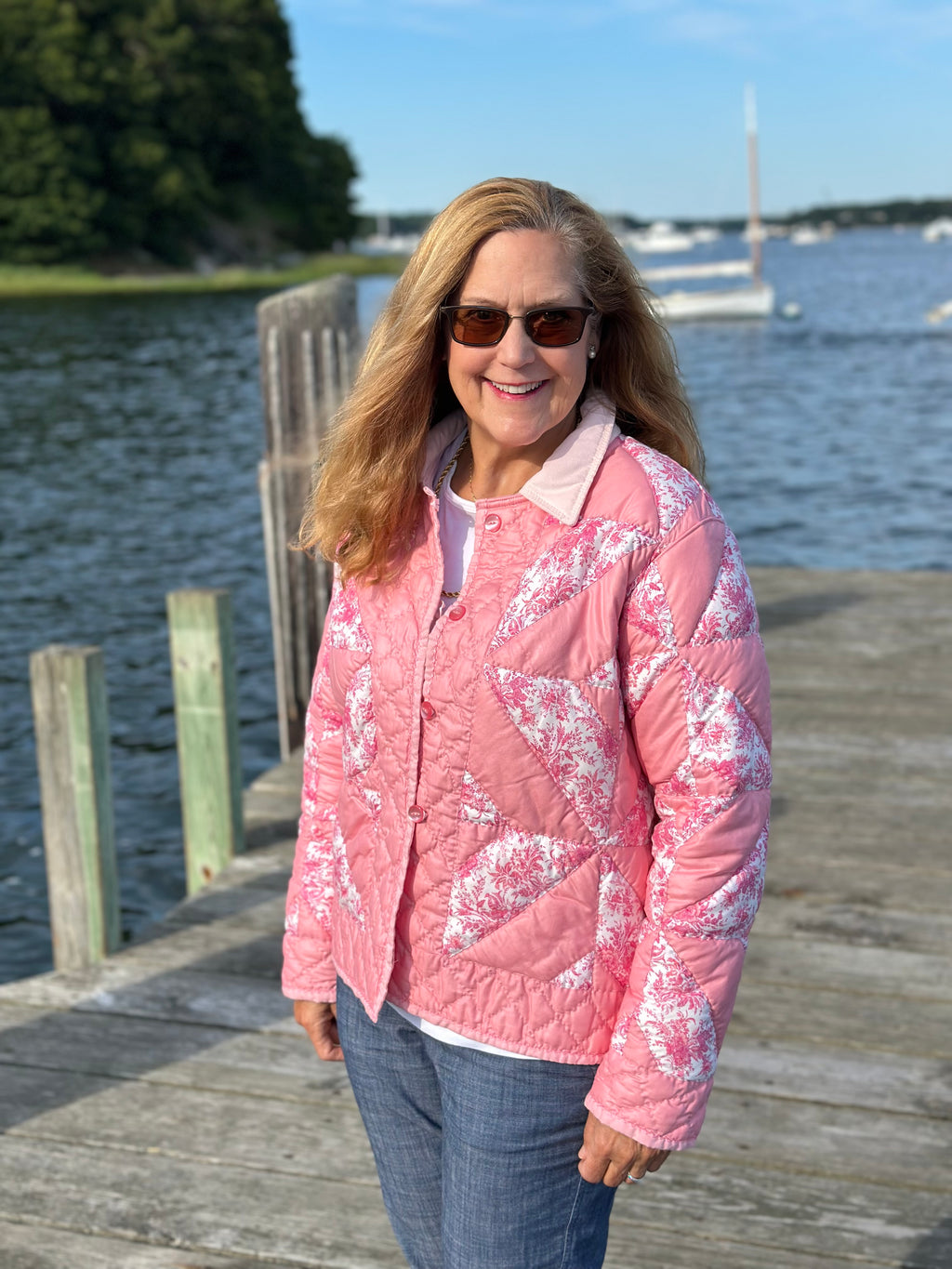 A woman wearing a pink quilted jacket with a white floral pattern stands on a wooden dock by the water, wearing blue jeans & sunglasses.