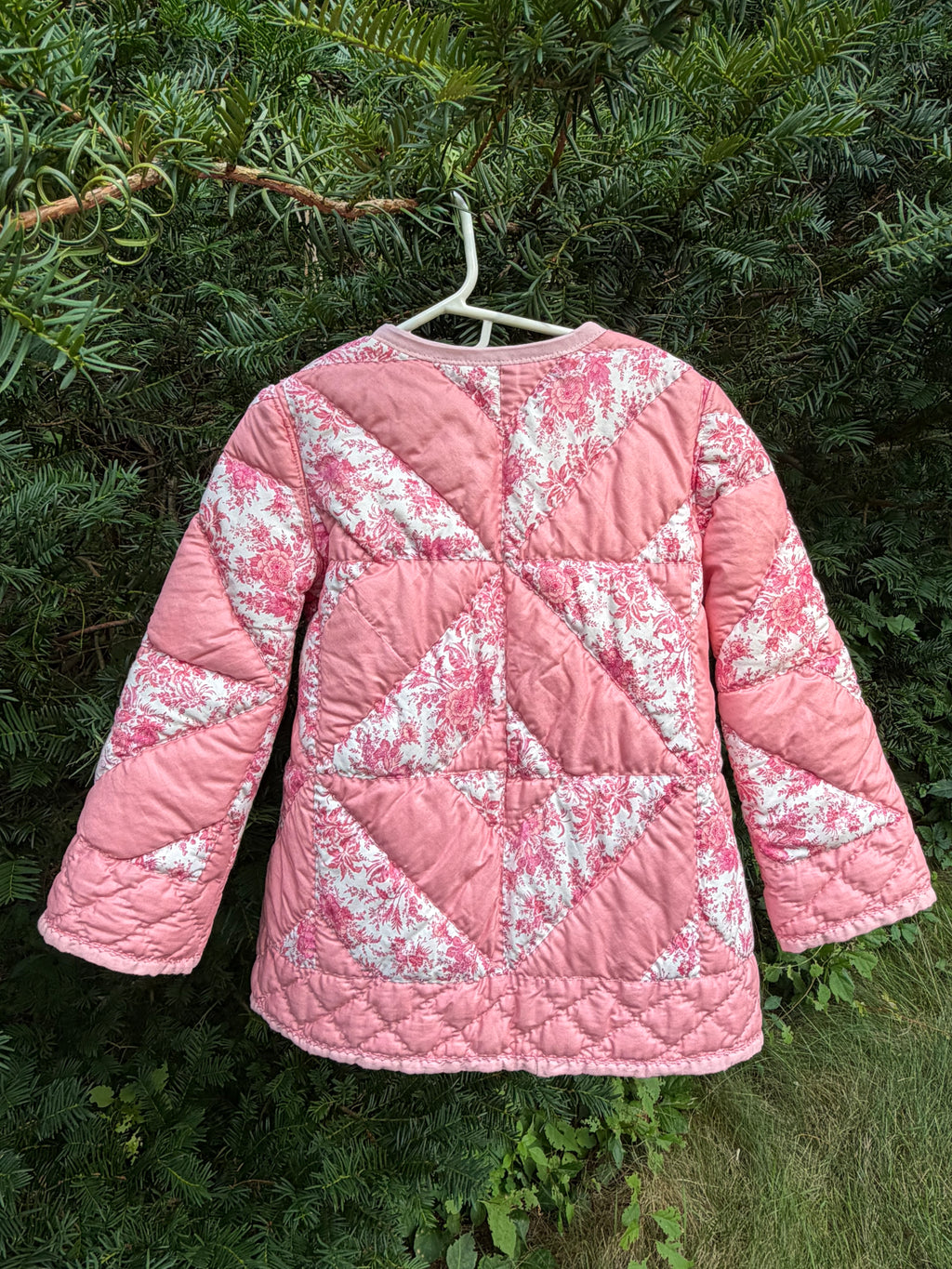 Pink handmade quilted jacket with a floral pattern, hanging on a white hanger against a background of green foliage.
